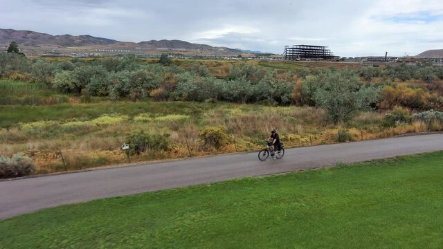 Senior man riding a bike along a paved path by a river with mountains and overcast sky - aerial follow view