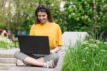 A beautiful and young Caucasian girl is sitting in nature with a laptop. Working, studying