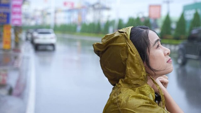 Asian girl wear yellow raincoat on the raining day standing on the road side, rainy season weather climate pouring rain, bad luck get wet while going out for work, car and motorcycle passing by
