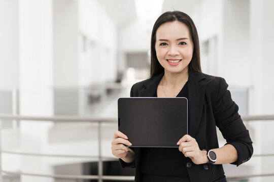 Portrait stock photo of a confident cheerful Asian businesswoman professional in black business suit using a digital tablet standing in the office building representing business oriented concept