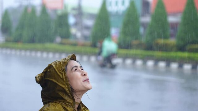 An asian girl with positive face expression on the pouring rain day, standing on the road side wearing yellow raincoat soaking wet in the rain, its rainy season in Asia, enjoying her day with rain