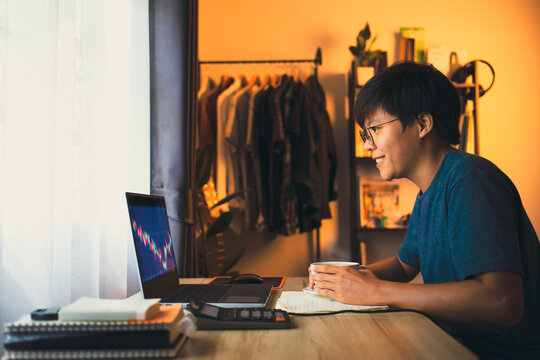 Asian young man 30s looking stock tickers or graphs cryptocurrency trading platforms on the laptop holding coffee ready start to hard work. work from home, financial at the home office in the morning - Powered by Adobe