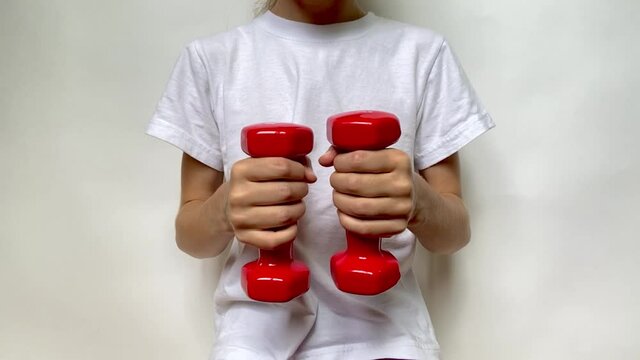 A girl in a white T-shirt slowly moves two red dumbbells weighing 1 kg 