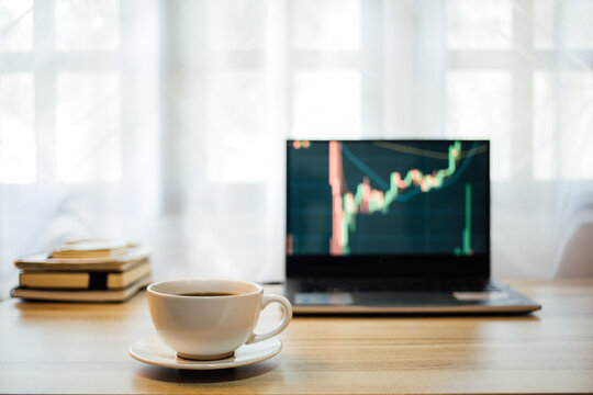 coffee Cup place table with stock tickers or graphs cryptocurrency trading platforms on the digital laptop device and a book, notebook on desktop at home office in the morning