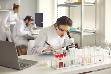 Female scientist doing research in a medical laboratory. Focused woman in goggles and latex gloves sits at a table in the lab and examines the cells under a microscope. Science and medicine concept.