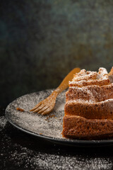 Top view of piece of yogurt sponge cake on dark plate with sugar and wooden fork, selective focus, on wooden table and gray background, vertical