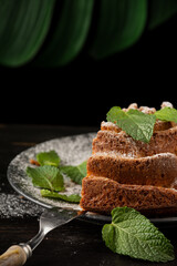Close-up of piece of yogurt sponge cake on plate with sugar, mint leaves and fork, selective focus, on wooden table and black background, vertical