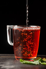 Close-up of glass with rooibos tea falling on wooden table with mint leaves, black background, vertical