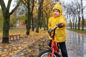 Portrait of boy in yellow raincoat on bicycle on autumn park background. Child riding bicycle in the rain