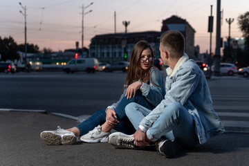 Fototapeta premium Modern young couple sit on the road on evening city background. Girl and a guy sort things out on the street.