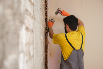 Young builder breaking up a house wall with a hammer and a chisel.