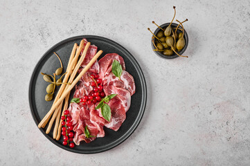 Slices of prosciutto coppa di parma ham with basil, grissini sticks, capers and redcurrant on dark...