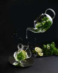 Floating glass tea pot with mint tea pouring into a transparent mug with lots of splashes.