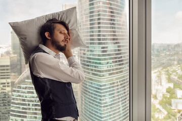 thoughtful businessman with a beard stands next to the window in the office against the backdrop of skyscrapers in Moscow in a white shirt and vest