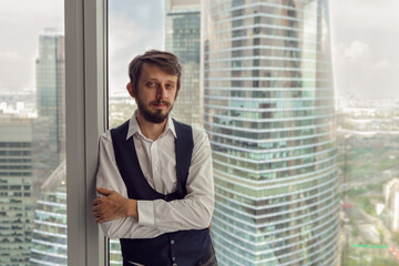 thoughtful businessman with a beard stands next to the window in the office against the backdrop of skyscrapers in Moscow in a white shirt and vest