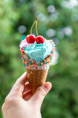 Female hand holds multi-colored ice cream with cherries in a waffle cone against blur green background