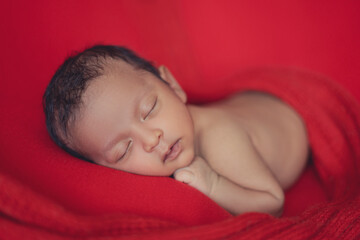 Obraz na płótnie Canvas newborn baby boy sleeping on stomach on red background. new born portrait. black hair face towards camera.