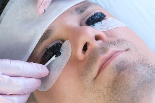 Beautician Is Wiping Black Paint From Man's Eyelashes On Laminating Lash Procedure. Cosmetologist Is Making Lash Lifting Lamination In Beauty Salon, Cosmetology Clinic, Closeup Face Of Client.