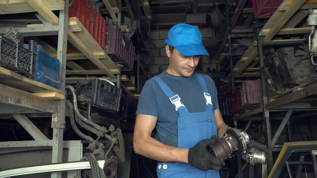 young Caucasian, employee of auto assembly, completes or checks car spare parts for client at auto parts warehouse. man in blue uniform. turns turbine from an internal combustion engine in his hands.