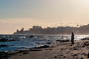 A fisherman with a fishing rod stands on the seashore with waves and small rocks against the backdrop of a coastal city during sunset