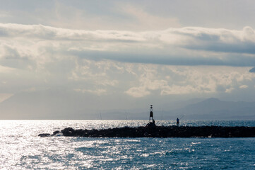 Two fishermen with fishing rods and a construction stand on a stone pier in the sea against the silhouette of mountains and clouds during sunset.