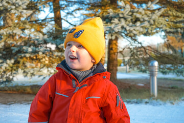 Smiling boy in red overalls and yellow hat walks in the yard in winter