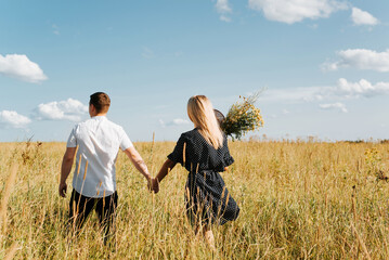 Happy young couple holding hands and walking in field, boyfriend and girlfriend in summer dress with bouquet of wild flowers, back view. Beautiful people in love enjoying time together. Love concept