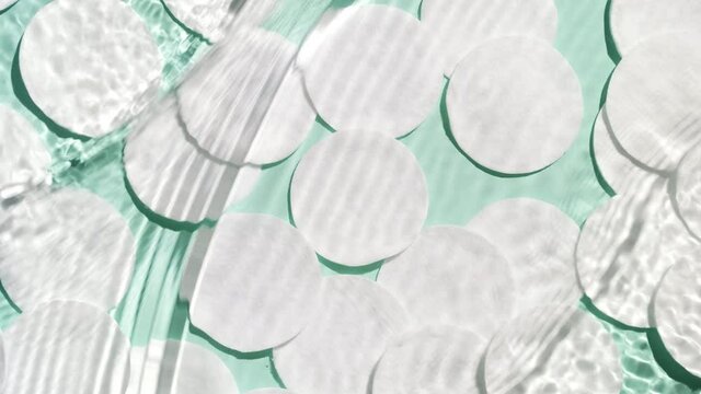 Multiple waves of water cross each other flowing from opposite sides and creating ripples spontaneously on pale green background with cotton pads | Background shot for face care products commercial