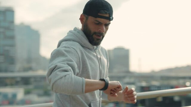 Bearded man in sport cap and hoodie spending morning time for outdoors activity. Male athlete running alone and checks the heart rate with a smart watch on city street. Healthy lifestyles.