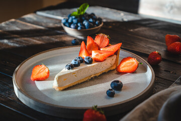 Slice of delicious cheesecake with strawberries and  blueberries on a ceramic plate on wooden vintage table. Dessert with juicy blueberries and strawberries close-up view.