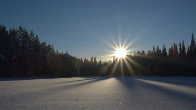 Time lapse of rising sun slowly moving horizontally behind spruce trees on a winter morning. Beautiful patterns from shadows on ice and diamond dust floating in the air.