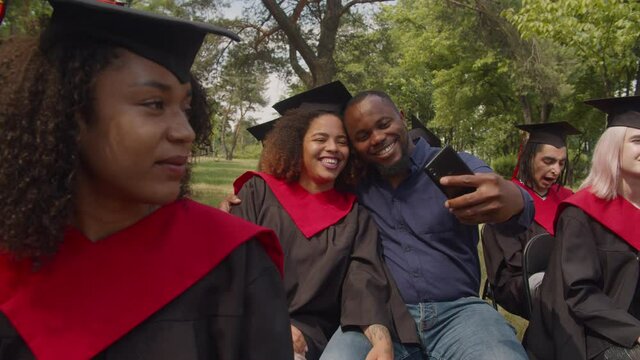 Carefree proud african father and joyful pretty african american daughter in graduation gown and mortarboard taking selfie shot on smart phone, expressing cheerful mood and happiness at graduation day