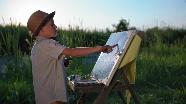 childhood hobby,talented little artist in hat draws with hands and paints on canvas with an easel near river backdrop of setting sun