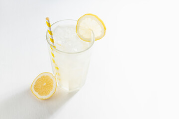 Homemade traditional refreshing cold sour lemonade infused beverage with ice, straw in glass with yellow lemon slice in sun beam on white wood table.