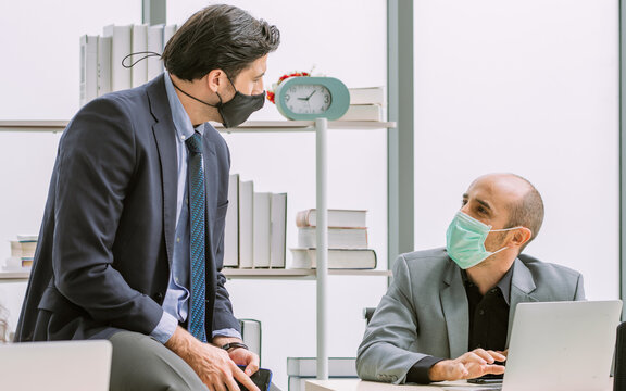 Two adult smart Caucasian business men wearing formal suit, working in indoor modern office, wearing face mask to protect virus for new normal and social distancing, meeting, discussing and talking.