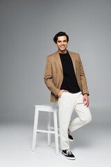 full length of happy young man in blazer standing with hand in pocket near white chair on grey