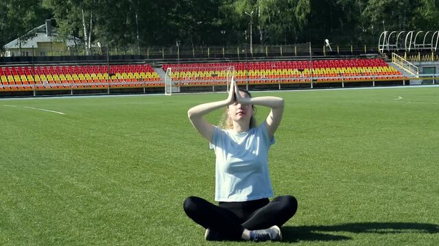 Young woman wearing blue T-shirt and black leggings is meditating after training at the stadium on a sunny day