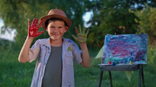 portrait of cheerful child with multi-colored palms in paints background of an easel with drawing on, smiles and looks at camera during summer vacation outdoor