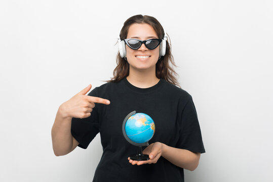 Photo of cool young woman, wearing sunglasses, headphones, pointing at globus and smiling at camera