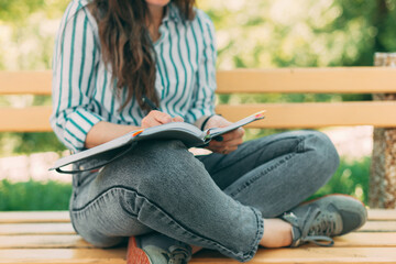 Photo of female with notebook and pen, sitting in a park on a bench. Woman writing in a notebook.