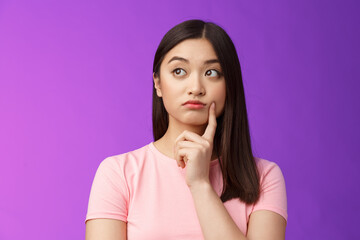 Close-up thoughtful cute asian girl dreamy standing purple background, touch cheek, turn left,...