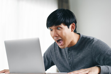 Shocked surprised Asian man looking at laptop computer screen sitting in the living room.