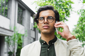 young man in eyeglasses and bomber jacket looking away while talking on smartphone outside
