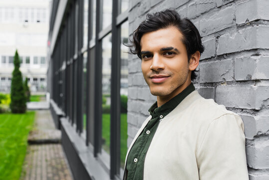 Cheerful Young Man In Bomber Jacket Smiling Near Brick Wall