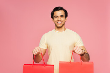 happy young man holding red shopping bags isolated on pink