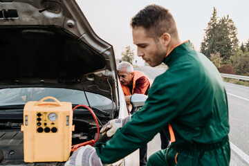 Two road assistant workers in towing service trying to start car engine with jump starter and...