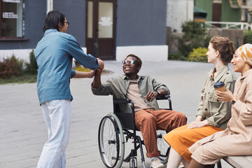 Group of young multi-ethnic friends including disabled man in wheelchair gathering together outdoors, men making handshake