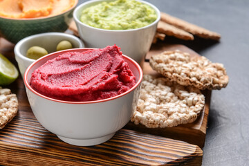 Bowls with tasty beet hummus and rice crackers on dark background, closeup