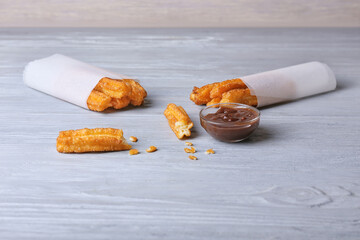 Tasty churros in parchment and melted chocolate sauce on color wooden background