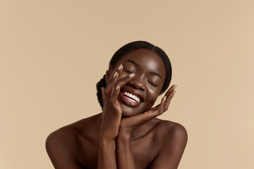Portrait close up of beautiful african girl with closed eyes. Smiling young woman touch her clean face. Concept of face skin care. Isolated on beige background. Studio shoot - obrazy, fototapety, plakaty